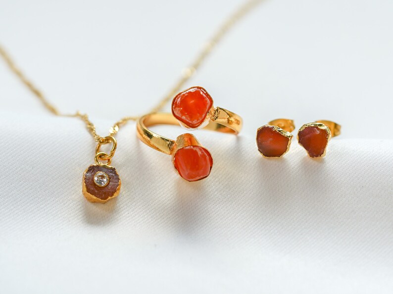 May include: A set of gold jewelry featuring three pieces with orange carnelian stones. The set includes a ring with two stones, a pair of earrings, and a pendant necklace.