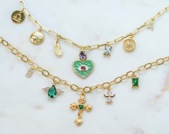 May include: Two gold chain necklaces with multiple charms. The charms include a green heart with an evil eye, a green teardrop with wings, a green square, a gold cross with a green stone, a gold sun, a gold feather, a gold zodiac symbol, a gold angel, and a blue gemstone.