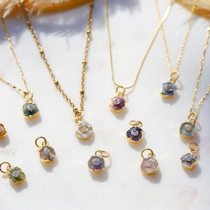 Könnte beinhalten: Eine Sammlung von goldenen Halsketten mit kleinen, bunten Edelsteinanhängern. Jeder Anhänger hat einen einzelnen, klaren Diamanten in der Mitte. Die Halsketten werden auf einer weißen Oberfläche präsentiert.