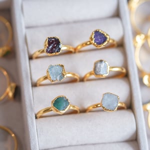 May include: A collection of gold-toned rings with various raw gemstone settings. The gemstones are in shades of purple, blue, green, and clear. The rings are displayed in a gray jewelry box, with additional gold rings visible.
