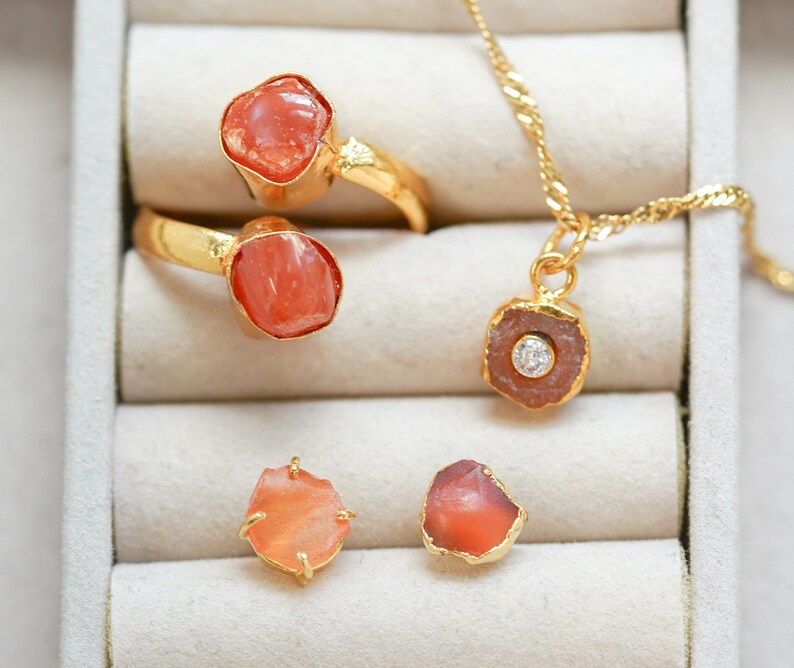 May include: Four pieces of jewelry featuring orange carnelian stones and gold settings. Two rings, a pendant, and a pair of earrings. The pendant has a small clear stone in the center.