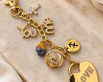May include: A gold-toned keychain featuring a lobster clasp, a crystal-encrusted cross, the words you and me in script, a raw blue stone, a circular lion charm, a Sagittarius zodiac symbol, and a heart-shaped padlock with the word LOVE.