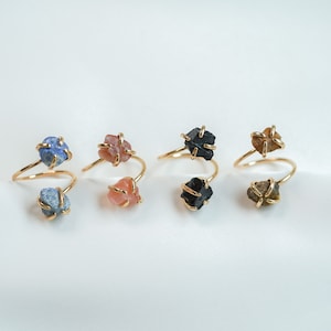May include: Five gold rings with different colored stones. The rings are made of a thin gold wire that is twisted around the stones. The stones are a light blue, a light pink, a black, a light brown, and a dark brown.