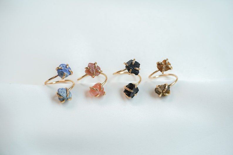 May include: Five gold rings with different colored stones. The rings are made of a thin gold wire that is twisted around the stones. The stones are a light blue, a light pink, a black, a light brown, and a dark brown.