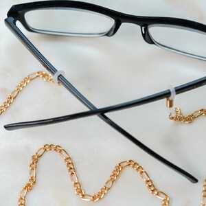 May include: Black eyeglasses with a gold chain attached to the arms. The chain is a curb chain style.