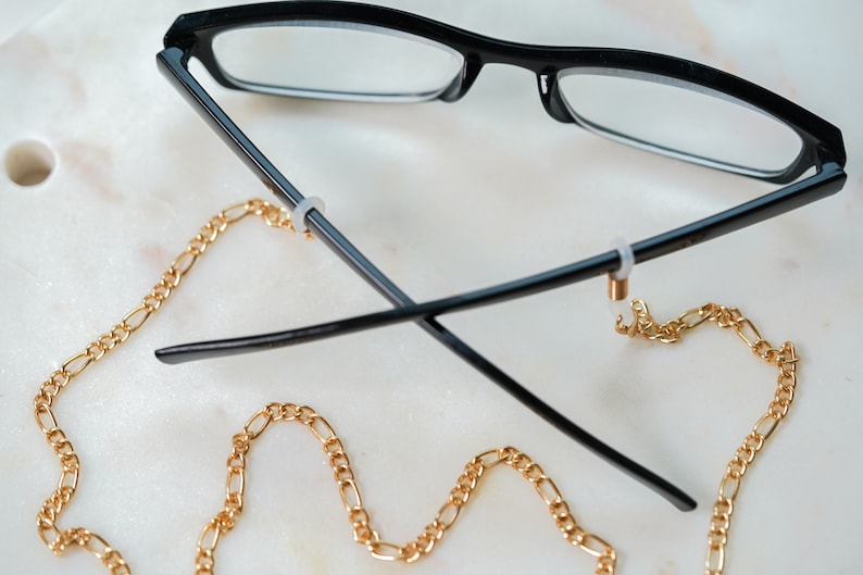 May include: Black eyeglasses with a gold chain attached to the arms. The chain is a curb chain style.