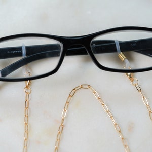 May include: Black framed glasses with a gold chain attached to the arms. The chain is made of oval links.