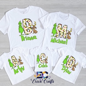 May include: White t-shirts with Grinch-themed designs. Each shirt features a large letter, a Grinch character, and a reindeer. Names are printed below the letters in green. The shirts are for a family.