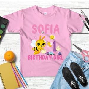 Pu&ograve; includere: Maglietta rosa con il nome "SOFIA", un'ape dei cartoni animati e la scritta "BIRTHDAY GIRL". &Egrave; presente anche il numero "1". L'immagine include materiali artistici, un'arancia e scarpe da ginnastica nere.