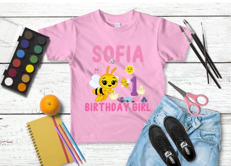 Pu&ograve; includere: Maglietta rosa con il nome "SOFIA", un'ape dei cartoni animati e la scritta "BIRTHDAY GIRL". &Egrave; presente anche il numero "1". L'immagine include materiali artistici, un'arancia e scarpe da ginnastica nere.