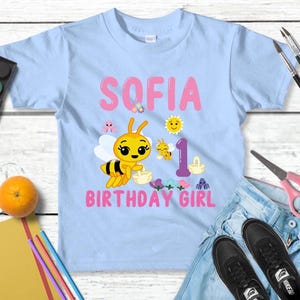 Pu&ograve; includere: Maglietta azzurra per bambini piccoli con il nome "SOFIA" e "BIRTHDAY GIRL" in rosa. Un'ape dei cartoni animati e altre grafiche decorano la maglietta. &Egrave; presente anche il numero "1". La maglietta &egrave; esposta con jeans, scarpe e materiale artistico.