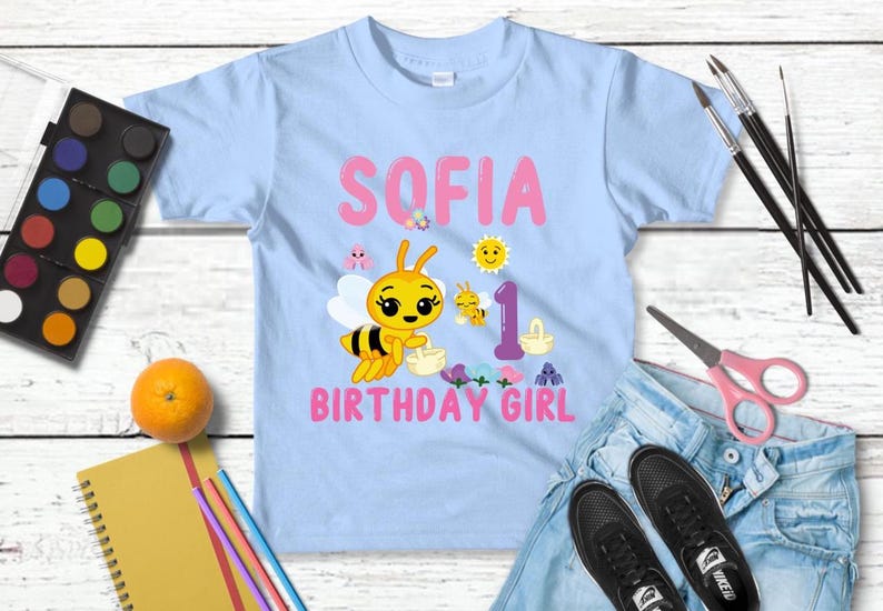 Pu&ograve; includere: Maglietta azzurra per bambini piccoli con il nome "SOFIA" e "BIRTHDAY GIRL" in rosa. Un'ape dei cartoni animati e altre grafiche decorano la maglietta. &Egrave; presente anche il numero "1". La maglietta &egrave; esposta con jeans, scarpe e materiale artistico.