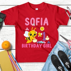 Pu&ograve; includere: Una maglietta rossa con il nome "SOFIA" in lettere rosa e una grafica di un'ape dei cartoni animati. La maglietta riporta anche la scritta "BIRTHDAY GIRL" in rosa. Anche il numero "1" &egrave; sulla maglietta. La maglietta &egrave; su una superficie di legno bianco.