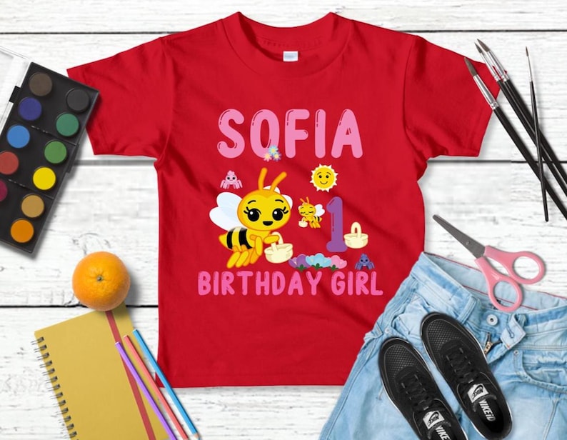 Pu&ograve; includere: Una maglietta rossa con il nome "SOFIA" in lettere rosa e una grafica di un'ape dei cartoni animati. La maglietta riporta anche la scritta "BIRTHDAY GIRL" in rosa. Anche il numero "1" &egrave; sulla maglietta. La maglietta &egrave; su una superficie di legno bianco.