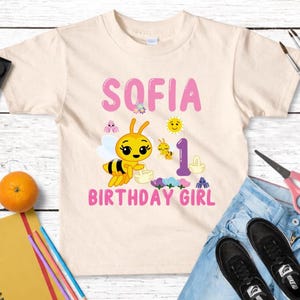 Pu&ograve; includere: Una maglietta beige chiaro per bambini con il nome "SOFIA" in lettere rosa e una grafica a forma di ape. La maglietta presenta anche la scritta "1 BIRTHDAY GIRL" ed &egrave; circondata da materiale artistico, jeans e scarpe.