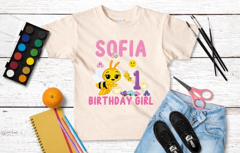 Pu&ograve; includere: Una maglietta beige chiaro per bambini con il nome "SOFIA" in lettere rosa e una grafica a forma di ape. La maglietta presenta anche la scritta "1 BIRTHDAY GIRL" ed &egrave; circondata da materiale artistico, jeans e scarpe.
