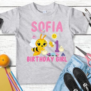 Pu&ograve; includere: Una maglietta grigio chiaro con il nome "SOFIA" in lettere rosa e una grafica di un'ape dei cartoni animati. La maglietta presenta anche le parole "BIRTHDAY GIRL" e il numero "1". Gli accessori includono forniture per la pittura, un'arancia e un paio di jeans.