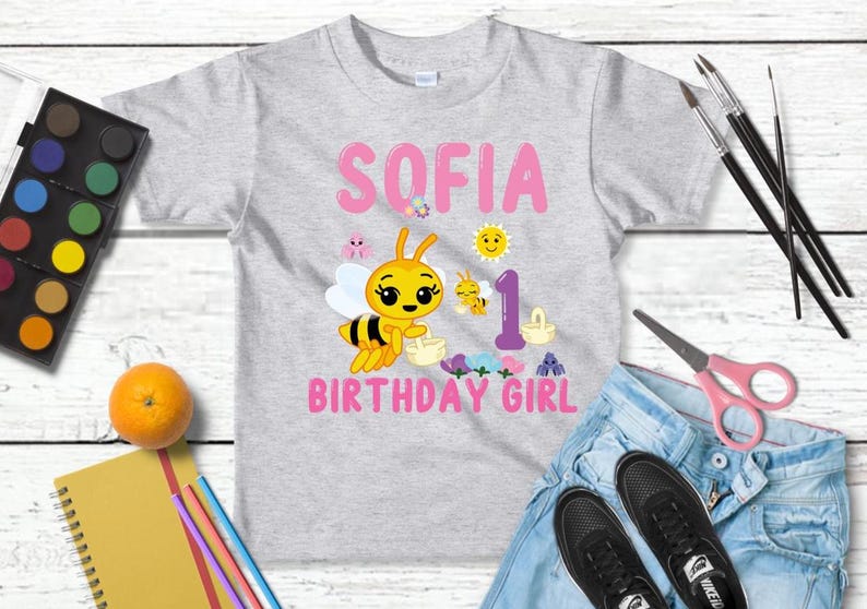 Pu&ograve; includere: Una maglietta grigio chiaro con il nome "SOFIA" in lettere rosa e una grafica di un'ape dei cartoni animati. La maglietta presenta anche le parole "BIRTHDAY GIRL" e il numero "1". Gli accessori includono forniture per la pittura, un'arancia e un paio di jeans.