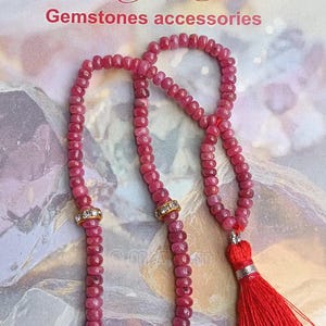 Könnte beinhalten: Eine Kette aus rosa Edelsteinperlen mit zwei goldfarbenen Zwischenelementen mit klaren Kristallen und einer leuchtend roten Quaste am Ende. Der Hintergrund zeigt den Text Pairking Gems Gemstones accessories in einer dekorativen Schrift.
