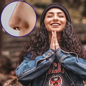 May include: A woman with curly brown hair is wearing a black beanie, a blue denim jacket, and a black shirt with a red and white logo. She is smiling and has her hands clasped in front of her. She has a nose ring.