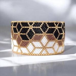 Op de afbeelding: Een brede armband gemaakt van kleine glaskralen in een geometrisch patroon. Het ontwerp bevat horizontale banden van matzwarte, metallic bronskleurige en iriserend witte kralen, gescheiden door rijen glanzende gouden kralen.