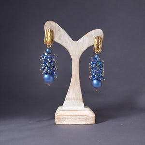 Può includere: Un paio di orecchini blu con perline e dettagli dorati. Gli orecchini sono esposti su un espositore di gioielli in legno chiaro su sfondo grigio. Ogni orecchino presenta un gruppo di piccole perline e una grande perla rotonda.