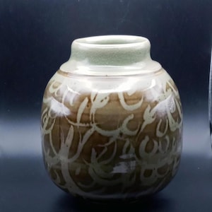 May include: A small, round ceramic vase with a light green glaze and a brown, abstract design. The vase has a wide mouth and a narrow base.