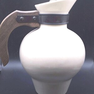 May include: A cream-colored ceramic pitcher with a rounded body and a tapered base. The pitcher features a dark brown wooden handle attached to the neck with a dark metal band secured by two visible screws. The spout is angled upward.