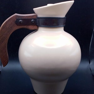 May include: A cream-colored ceramic pitcher with a wooden handle and a black metal band around the neck.