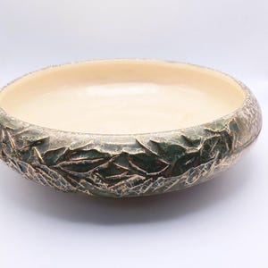 May include: A shallow ceramic bowl with a smooth, cream-colored interior and a textured exterior featuring a dark green glaze with a prominent gold-toned crackle pattern. The bowl has a rounded rim and a wide, circular base, presenting a distinct contrast between the light inner surface and the dark, weathered-looking outer finish.
