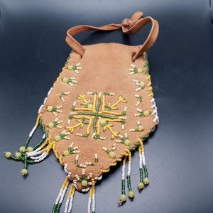 May include: A brown leather pouch with a beaded design. The pouch is decorated with white, green, and yellow beads. The pouch has a drawstring closure and a fringe.
