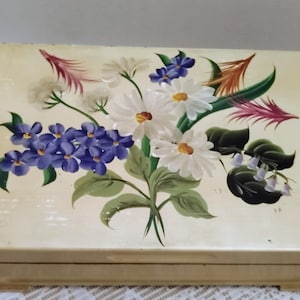 May include: A rectangular, cream-colored box with a hand-painted floral design. The artwork features white daisies, purple violets, and other flowers with green leaves. The box has a slightly distressed appearance, suggesting it may be vintage or antique.