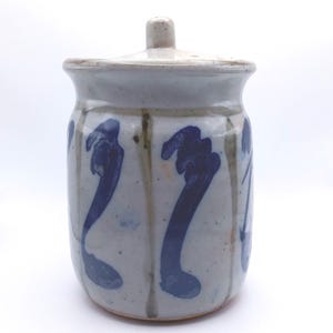 May include: A cylindrical ceramic jar with a matching lid, featuring a speckled light gray glaze and vertical, hand-painted dark blue brushstroke patterns. The jar has a slightly flared rim and a small, knob-like handle on the center of the lid.
