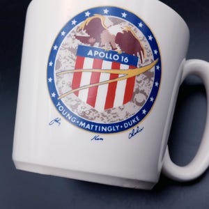 May include: White ceramic mug with a blue and red Apollo 16 mission patch featuring an eagle and the text "Apollo 16". The patch is surrounded by a circle of stars. The text "Young, Mattingly, Duke" is printed below the patch with the astronauts' signatures.