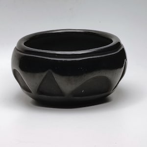 May include: A small, dark gray ceramic bowl with a rounded shape and a wide opening. The bowl features a series of carved, repeating triangular patterns around its exterior. The bowl is sitting on a white surface.