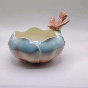 May include: A ceramic bowl with a bird accent. The bowl is a light blue and peach color with a scalloped rim and a peach-colored bird perched on the side. The interior is a cream color.