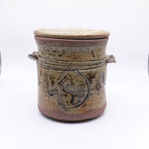 May include: A cylindrical ceramic jar with a matching lid, featuring a mottled tan and brown glaze. The vessel has two small side handles and a textured, hand-formed appearance with a dark, earthy base and a decorative incised circular pattern.