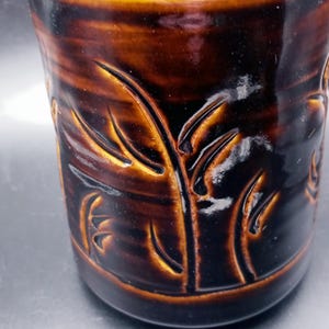 May include: A brown ceramic mug with a dark brown glaze and a carved design of leaves. The mug has a glossy finish.