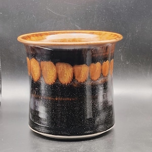 May include: A cylindrical ceramic vase with a glossy finish. The vase features a dark brown base, a band of orange-brown oval shapes, and a lighter brown rim. The interior is also brown. The vase has a speckled texture.