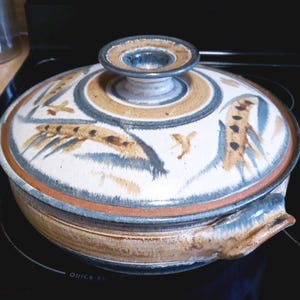 May include: A round ceramic casserole dish with a matching lid, featuring a hand-painted wheat motif in shades of tan, blue, and cream. The pot has a wide, flat base, a central knob handle on the lid, and integrated side handles on the main body.