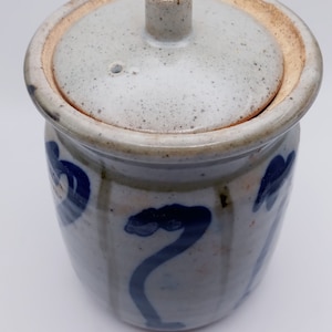May include: A white ceramic jar with a lid, decorated with blue floral designs. The jar has a textured surface and a slightly rounded shape.