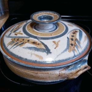 May include: A ceramic casserole dish with a lid. The dish has a white background with brown and blue painted designs. The lid has a knob on top.
