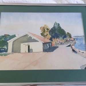 May include: A framed watercolor painting depicting a coastal scene with a light-colored building featuring a reddish roof, situated near a body of water. The landscape includes green trees, a paved area, and a signature in the bottom right corner.