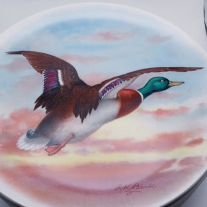 May include: A decorative plate featuring a flying duck with brown, white, green, and purple feathers. The duck is set against a backdrop of a sunset with pink, orange, and blue hues. The plate has a brown rim and is signed "R.K. Beck".