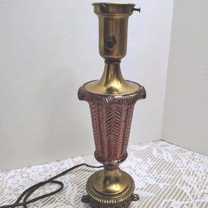 May include: A table lamp base featuring a brass socket and a central column with a textured, copper-colored glass body displaying a repeating leaf pattern. The base is constructed of ornate brass with a circular, footed design and a brown power cord.