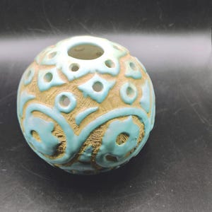 May include: A spherical ceramic vase featuring a textured tan surface with raised, glossy light blue floral and geometric patterns. The object has a circular opening at the top and several small, evenly spaced circular cutouts throughout.