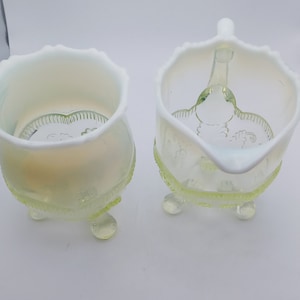 May include: A pair of antique, opalescent glass serving pieces. The set includes a creamer with a handle and a sugar bowl, both with a scalloped rim and resting on three small feet. The glass has a subtle, iridescent sheen and a delicate, floral design.