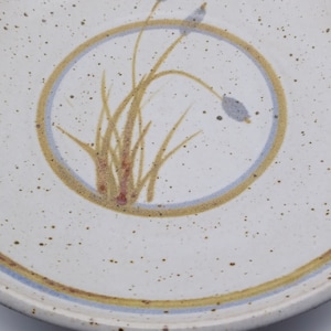 May include: A white ceramic plate with a brown and blue rim. The center of the plate features a circle with a brown and blue design of stylized grass.