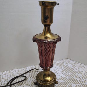 May include: A vintage table lamp with a brass base and accents. The lamp features a burgundy-colored, textured glass shade with a woven pattern. The lamp is approximately 12 inches tall.