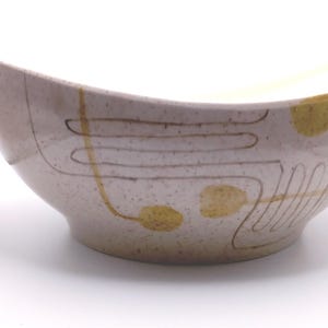 May include: A speckled off-white ceramic bowl featuring a curved, elongated shape with two handles. The exterior is decorated with thin, dark brown linear patterns and solid mustard yellow circular accents arranged in a flowing, abstract design.